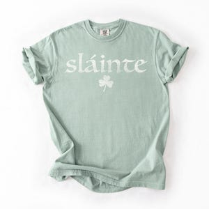 May include: A light green t-shirt with the word "sl&aacute;inte" in white, along with a white shamrock. The shirt has rolled-up sleeves and a crew neck. The shirt is made of a soft material.