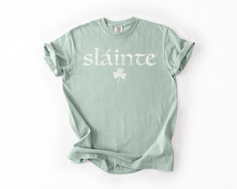 May include: A light green t-shirt with the word "sl&aacute;inte" in white, along with a white shamrock. The shirt has rolled-up sleeves and a crew neck. The shirt is made of a soft material.