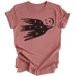 Peut inclure: Un t-shirt en mauve chin&eacute; avec un motif noir d'un oiseau stylis&eacute; volant avec des &eacute;toiles et une lune croissante.