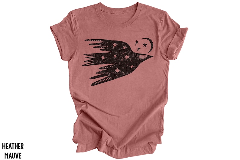 Peut inclure: Un t-shirt en mauve chin&eacute; avec un motif noir d'un oiseau stylis&eacute; volant avec des &eacute;toiles et une lune croissante.