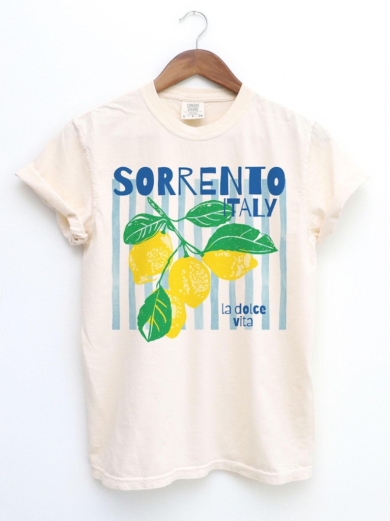 Puede incluir: Camiseta color crema con las palabras "SORRENTO ITALY" en letras azules. Debajo, un gr&aacute;fico de limones y hojas, con "la dolce vita" impreso debajo. La camiseta est&aacute; colgada en una percha de madera. El dise&ntilde;o tiene rayas verticales azules.