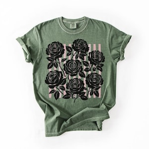 May include: Olive green t-shirt featuring a graphic of black roses on a pink striped background. The roses are detailed with stems and leaves. The shirt has a classic crew neck and rolled sleeves.