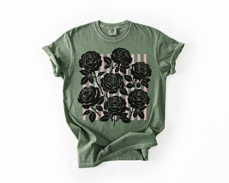 May include: Olive green t-shirt featuring a graphic of black roses on a pink striped background. The roses are detailed with stems and leaves. The shirt has a classic crew neck and rolled sleeves.