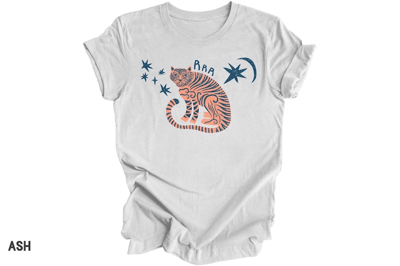 Peut inclure: Un t-shirt blanc avec une illustration de tigre de couleur p&ecirc;che et le mot "RAA" au-dessus. Le tigre est entour&eacute; d'&eacute;toiles bleues et d'un croissant de lune.