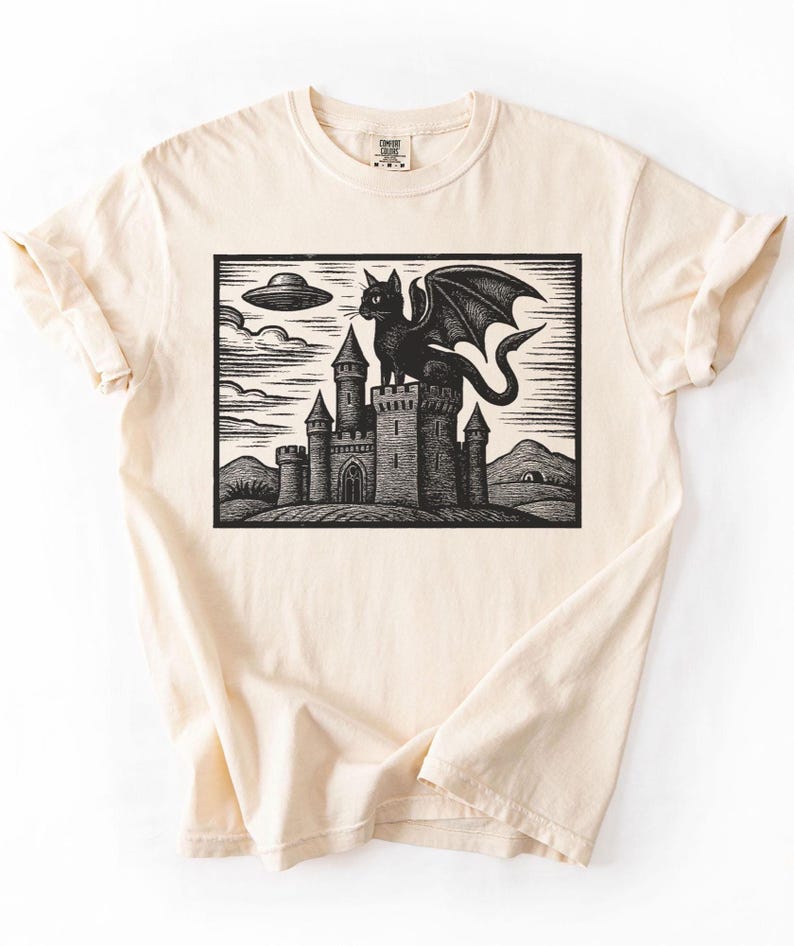 May include: Cream-colored t-shirt featuring a black and white graphic. The graphic depicts a castle with a winged cat on top, an alien spaceship, and a landscape. The shirt has short sleeves and a crew neck.