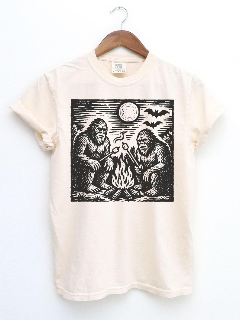 May include: Cream-colored t-shirt featuring a black and white graphic of two Bigfoot creatures roasting marshmallows over a campfire under a full moon. Bats are silhouetted in the sky above the moon. The shirt is hanging on a wooden hanger.