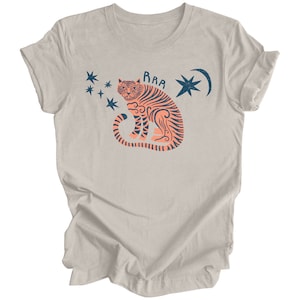Peut inclure: Un t-shirt couleur poussi&egrave;re de bruy&egrave;re avec un motif de tigre avec une lune croissante et des &eacute;toiles. Le texte "RAA" est au-dessus du tigre.