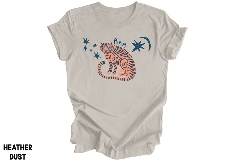 Peut inclure: Un t-shirt couleur poussi&egrave;re de bruy&egrave;re avec un motif de tigre avec une lune croissante et des &eacute;toiles. Le texte "RAA" est au-dessus du tigre.