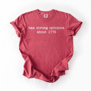 Puede incluir: Camiseta roja con el texto blanco "has strong opinions about 1776". La camiseta tiene mangas enrolladas y cuello redondo. La tela parece suave y c&oacute;moda.