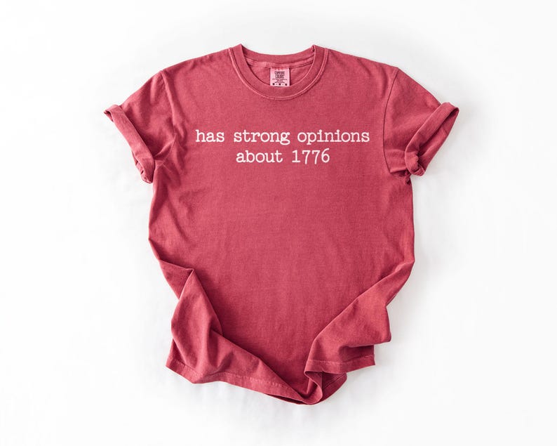 Puede incluir: Camiseta roja con el texto blanco "has strong opinions about 1776". La camiseta tiene mangas enrolladas y cuello redondo. La tela parece suave y c&oacute;moda.