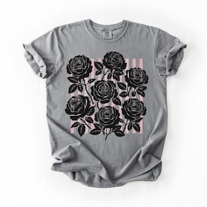 May include: Gray t-shirt featuring a black rose graphic on a pink striped background. The roses are detailed with stems and leaves. The shirt has a crew neck and short sleeves.
