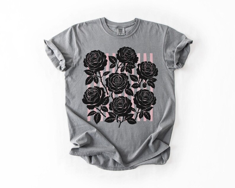 May include: Gray t-shirt featuring a black rose graphic on a pink striped background. The roses are detailed with stems and leaves. The shirt has a crew neck and short sleeves.
