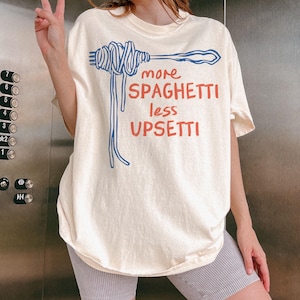 May include: A white t-shirt with a blue and red graphic of spaghetti on a fork. The text on the shirt reads "more spaghetti less upsetti".