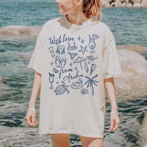 Puede incluir: Camiseta color crema con ilustraciones azules de elementos tem&aacute;ticos de playa, como un cangrejo, un bikini, chanclas y un c&oacute;ctel. El texto "With love from Aruba" tambi&eacute;n est&aacute; impreso en la camiseta.