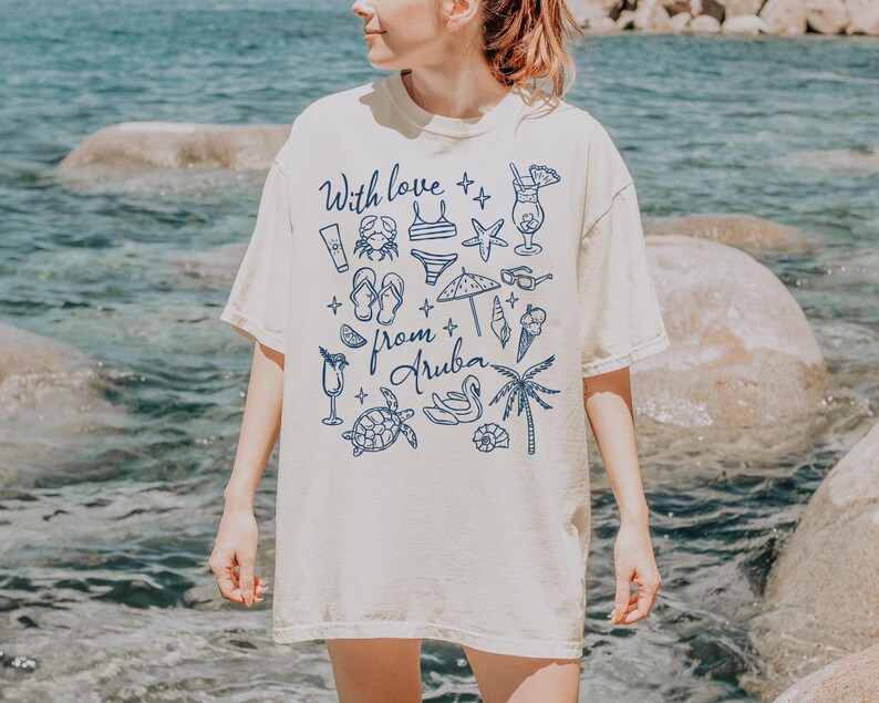 Puede incluir: Camiseta color crema con ilustraciones azules de elementos tem&aacute;ticos de playa, como un cangrejo, un bikini, chanclas y un c&oacute;ctel. El texto "With love from Aruba" tambi&eacute;n est&aacute; impreso en la camiseta.