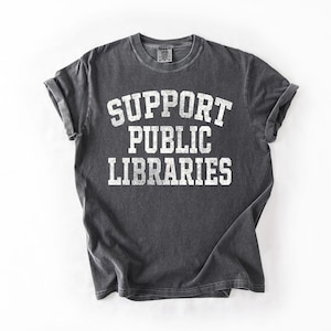 May include: A dark gray t-shirt with the words "SUPPORT PUBLIC LIBRARIES" printed in large, white, block letters. The shirt has rolled-up sleeves and a vintage, worn look.