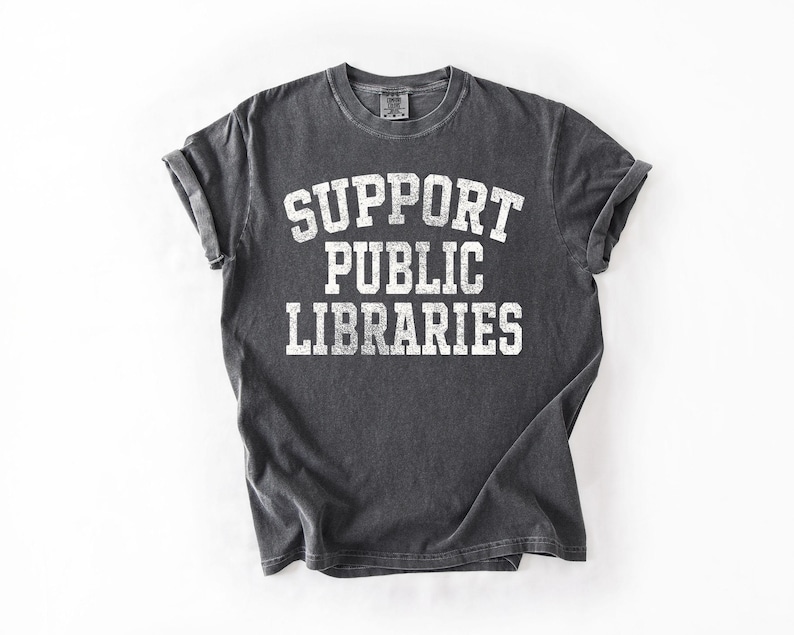 May include: A dark gray t-shirt with the words "SUPPORT PUBLIC LIBRARIES" printed in large, white, block letters. The shirt has rolled-up sleeves and a vintage, worn look.