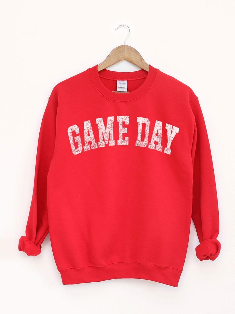 May include: A bright red sweatshirt with the text "GAME DAY" in distressed white lettering. The crew neck sweatshirt is hanging on a wooden hanger. The sleeves are cuffed at the wrists.