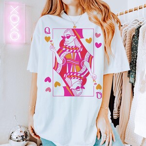 May include: White t-shirt featuring a Queen of Hearts playing card design in pink and gold. The graphic includes hearts and the letter "Q". The word "WHITE" is printed at the bottom. A neon sign with "XOXO" is in the background.