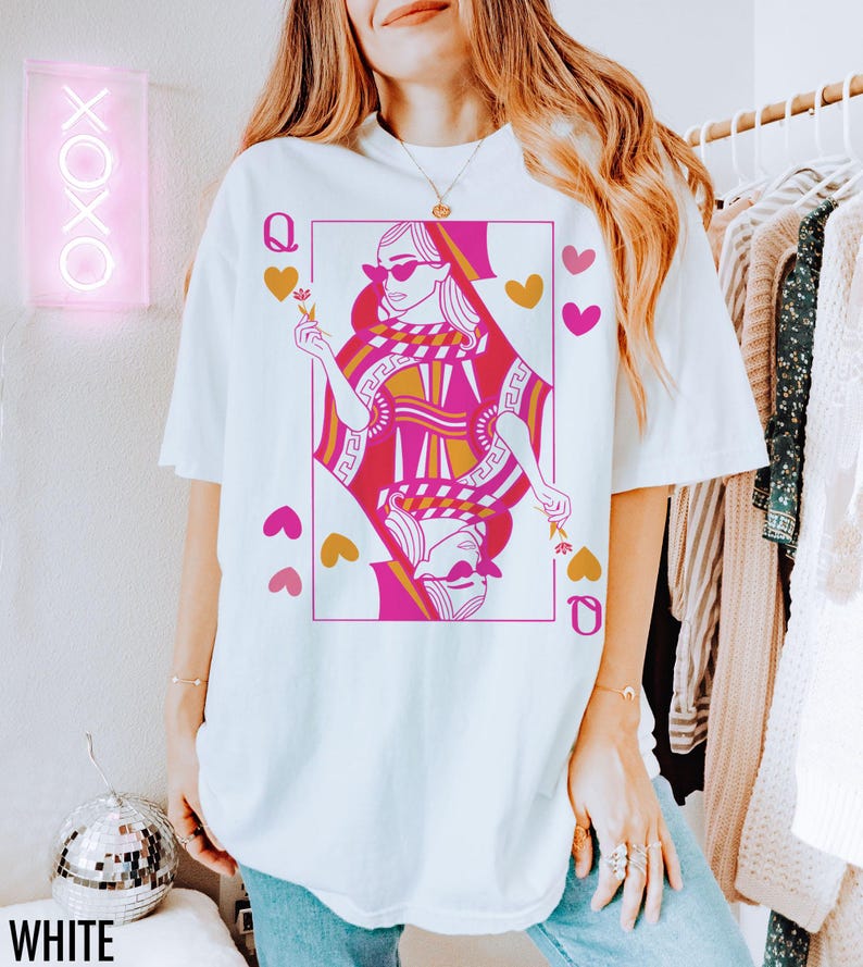 May include: White t-shirt featuring a Queen of Hearts playing card design in pink and gold. The graphic includes hearts and the letter "Q". The word "WHITE" is printed at the bottom. A neon sign with "XOXO" is in the background.