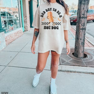 May include: A woman wearing a light beige oversized t-shirt with a graphic of a hot dog with the text "Bad Day To Be A Hot Dog". She is wearing white sneakers and standing on a sidewalk.