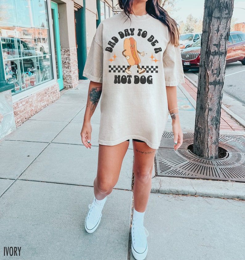 May include: A woman wearing a light beige oversized t-shirt with a graphic of a hot dog with the text "Bad Day To Be A Hot Dog". She is wearing white sneakers and standing on a sidewalk.