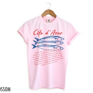 May include: A light pink t-shirt with the words "C&ocirc;te d'Azur" in red script above three blue fish illustrations. Below the fish is a paragraph of text in red script. The word "Blossom" is printed in black at the bottom left.