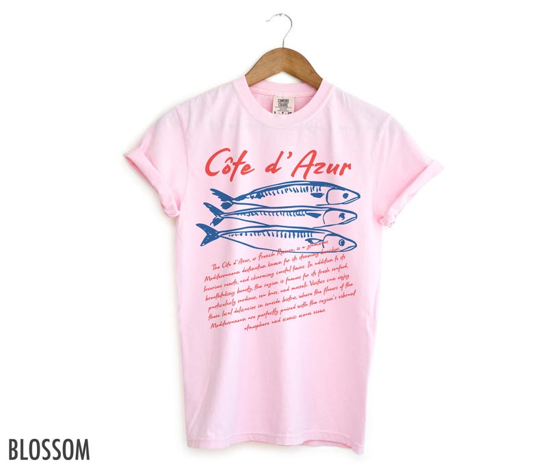 May include: A light pink t-shirt with the words "C&ocirc;te d'Azur" in red script above three blue fish illustrations. Below the fish is a paragraph of text in red script. The word "Blossom" is printed in black at the bottom left.