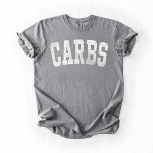 May include: A gray t-shirt with the word "CARBS" printed in large, white, block letters. The shirt is folded and has rolled-up sleeves. The fabric appears soft and slightly worn.