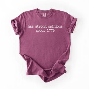 Puede incluir: Camiseta de color granate con el texto blanco "has strong opinions about 1776". La camiseta tiene mangas cortas y cuello redondo.