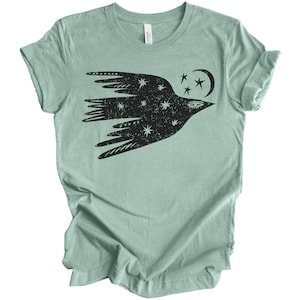 Peut inclure: Un t-shirt bleu clair avec un motif noir d'un oiseau stylis&eacute; volant avec des &eacute;toiles et une lune croissante. L'oiseau est rempli de petites &eacute;toiles blanches.