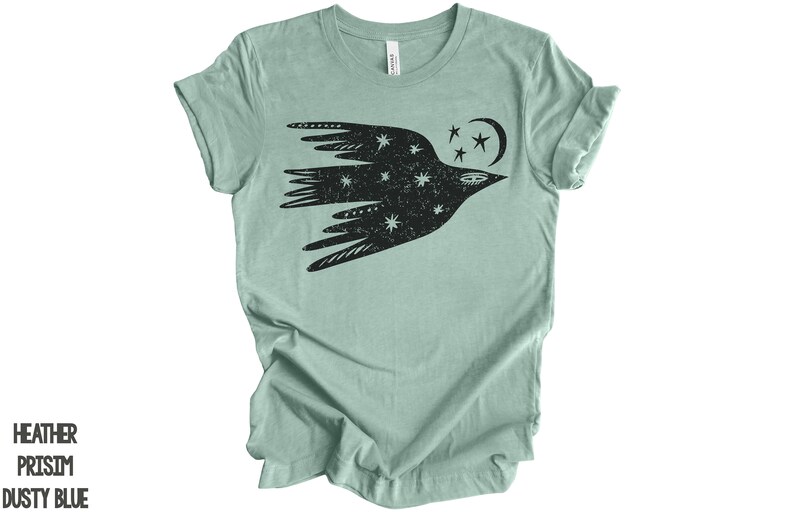 Peut inclure: Un t-shirt bleu clair avec un motif noir d'un oiseau stylis&eacute; volant avec des &eacute;toiles et une lune croissante. L'oiseau est rempli de petites &eacute;toiles blanches.