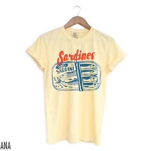 K&ouml;nnte beinhalten: Ein gelbes T-Shirt mit kurzen &Auml;rmeln und einem Aufdruck einer Sardinenb&uuml;chse. Der Text "Sardines" ist in Rot auf dem Shirt gedruckt. Die Dose tr&auml;gt den Text "WC Sardine".
