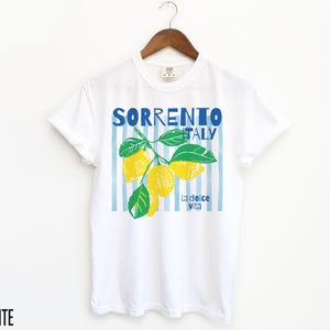 Puede incluir: Camiseta blanca con un dise&ntilde;o gr&aacute;fico de limones y el texto "SORRENTO ITALY" en azul. El dise&ntilde;o incluye hojas verdes y la frase "la dolce vita". La camiseta est&aacute; colgada de una percha de madera.