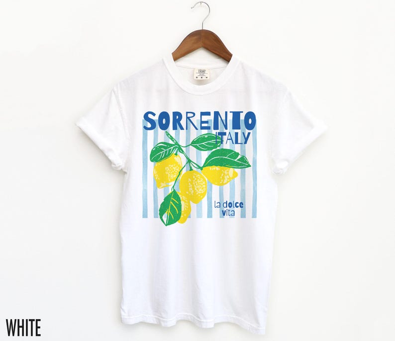 Puede incluir: Camiseta blanca con un dise&ntilde;o gr&aacute;fico de limones y el texto "SORRENTO ITALY" en azul. El dise&ntilde;o incluye hojas verdes y la frase "la dolce vita". La camiseta est&aacute; colgada de una percha de madera.