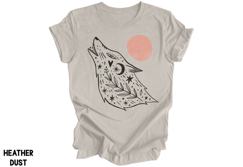 May include: A heather grey t-shirt featuring a black and white illustration of a wolf howling at a pink sun. The wolf is surrounded by stars, leaves, and other celestial symbols.