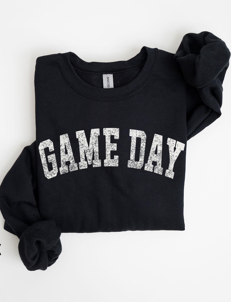 May include: Black sweatshirt with the text "GAME DAY" in distressed white lettering. The crew neck sweatshirt is folded, with the sleeves neatly tucked in. The Gildan brand tag is visible at the neckline. A casual, comfortable apparel item.