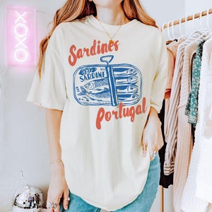 Op de afbeelding: Een wit T-shirt met een afbeelding van een blikje sardines met de tekst "Sardines Portugal". Het blikje heeft een blauw en wit ontwerp met een rood label waarop "Sardine" en "Portugese filets in olie" staat.