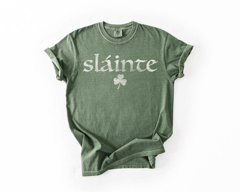 May include: Green t-shirt with the word "sl&aacute;inte" in a distressed white font and a small shamrock graphic. The shirt has a crew neck and rolled-up sleeves, and is made of a soft, comfortable material.