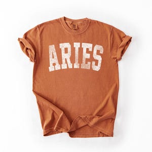 May include: A terracotta-colored t-shirt with the word "ARIES" printed in white, block letters. The shirt has a crew neck and rolled-up sleeves. The fabric has a slightly faded, vintage look.