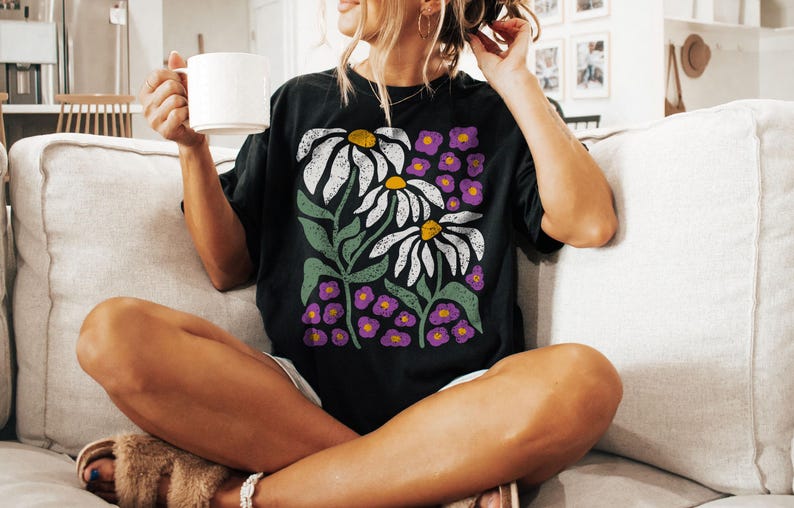May include: Black t-shirt featuring a floral design with white daisies, yellow centers, and purple accent flowers. The graphic tee has green stems and leaves. The person is holding a white mug.