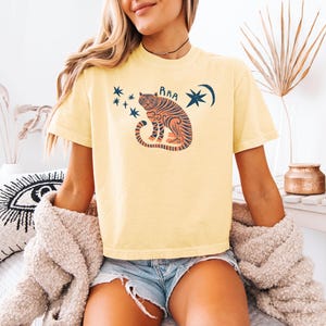 May include: Yellow t-shirt with a tiger graphic, stars, and a crescent moon. The tiger is orange and black with the word "RAA" above it. The shirt is worn with denim shorts and a beige cardigan.