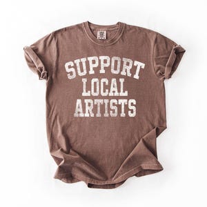 May include: A brown t-shirt with the text "SUPPORT LOCAL ARTISTS" in white, distressed lettering. The shirt has a crew neck and short sleeves, and is folded on a white surface.