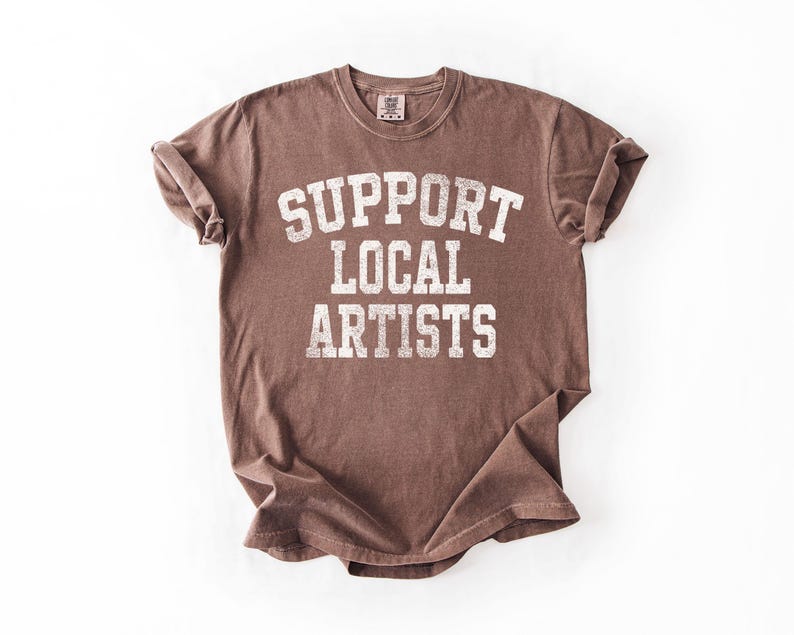 May include: A brown t-shirt with the text "SUPPORT LOCAL ARTISTS" in white, distressed lettering. The shirt has a crew neck and short sleeves, and is folded on a white surface.