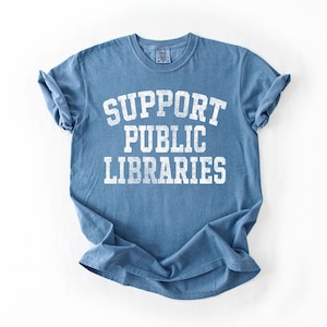May include: A blue t-shirt with the words "SUPPORT PUBLIC LIBRARIES" printed in white, distressed lettering. The shirt has short sleeves and a crew neck. The fabric is slightly wrinkled.