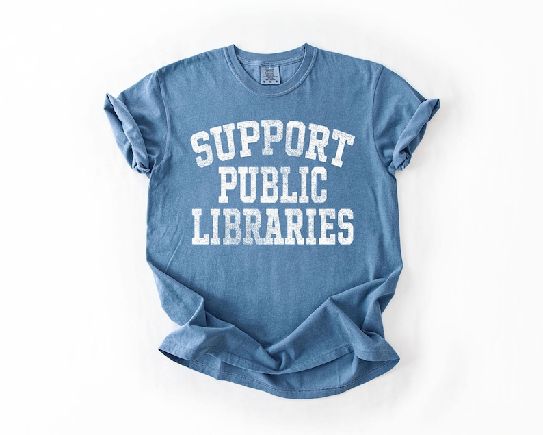 May include: A blue t-shirt with the words "SUPPORT PUBLIC LIBRARIES" printed in white, distressed lettering. The shirt has short sleeves and a crew neck. The fabric is slightly wrinkled.