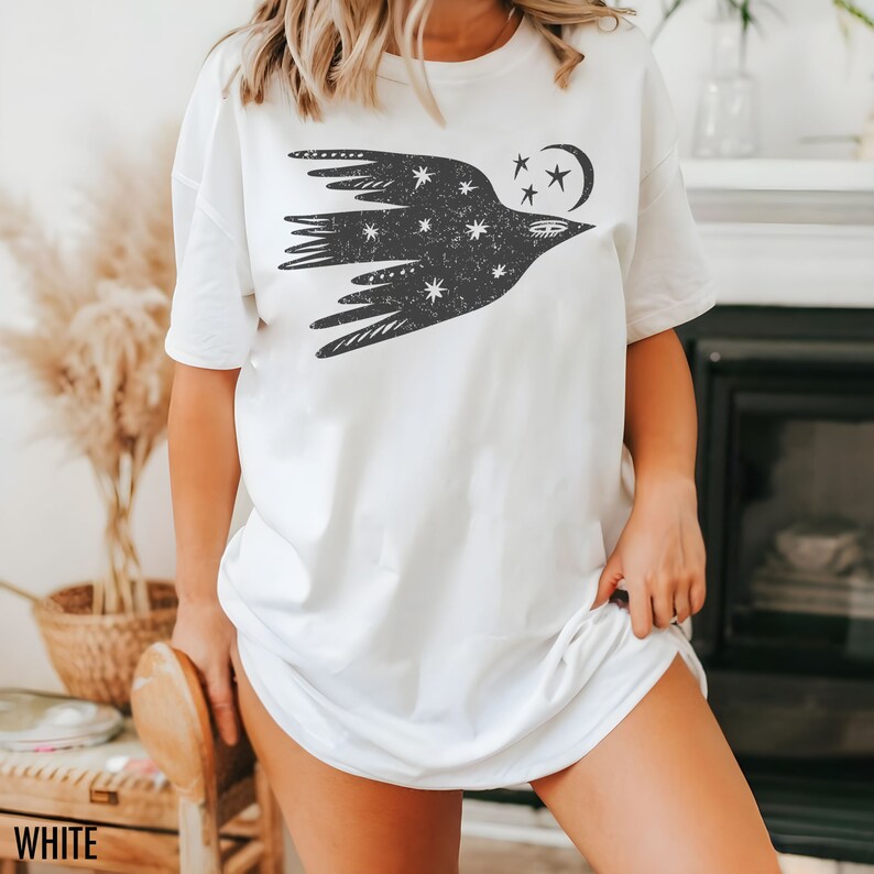May include: White t-shirt featuring a black graphic of a bird with stars and a crescent moon. The bird design is centered on the front of the shirt. The word "WHITE" is printed at the bottom left corner of the image.