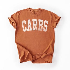 May include: A rust-colored t-shirt with the word "CARBS" printed in large, distressed white letters across the front. The shirt has short sleeves and a crew neck. The fabric appears soft and slightly faded.