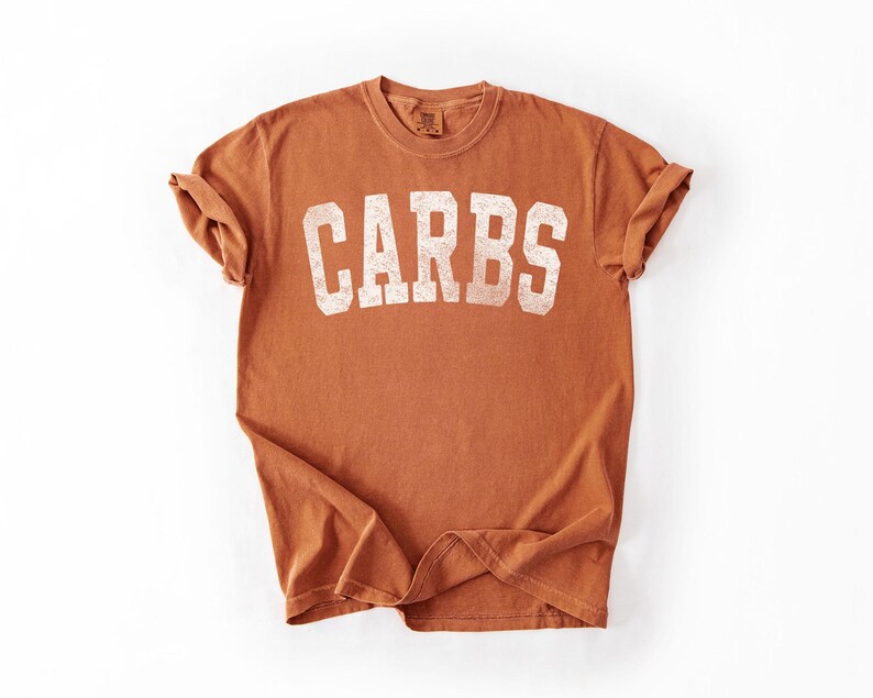 May include: A rust-colored t-shirt with the word "CARBS" printed in large, distressed white letters across the front. The shirt has short sleeves and a crew neck. The fabric appears soft and slightly faded.