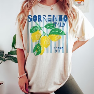 Puede incluir: Camiseta color crema con las palabras "SORRENTO ITALY" en azul, con un gr&aacute;fico de rama de lim&oacute;n. La camiseta tambi&eacute;n tiene la frase "la dolce vita" en azul. El dise&ntilde;o est&aacute; sobre un fondo de rayas verticales azul claro.